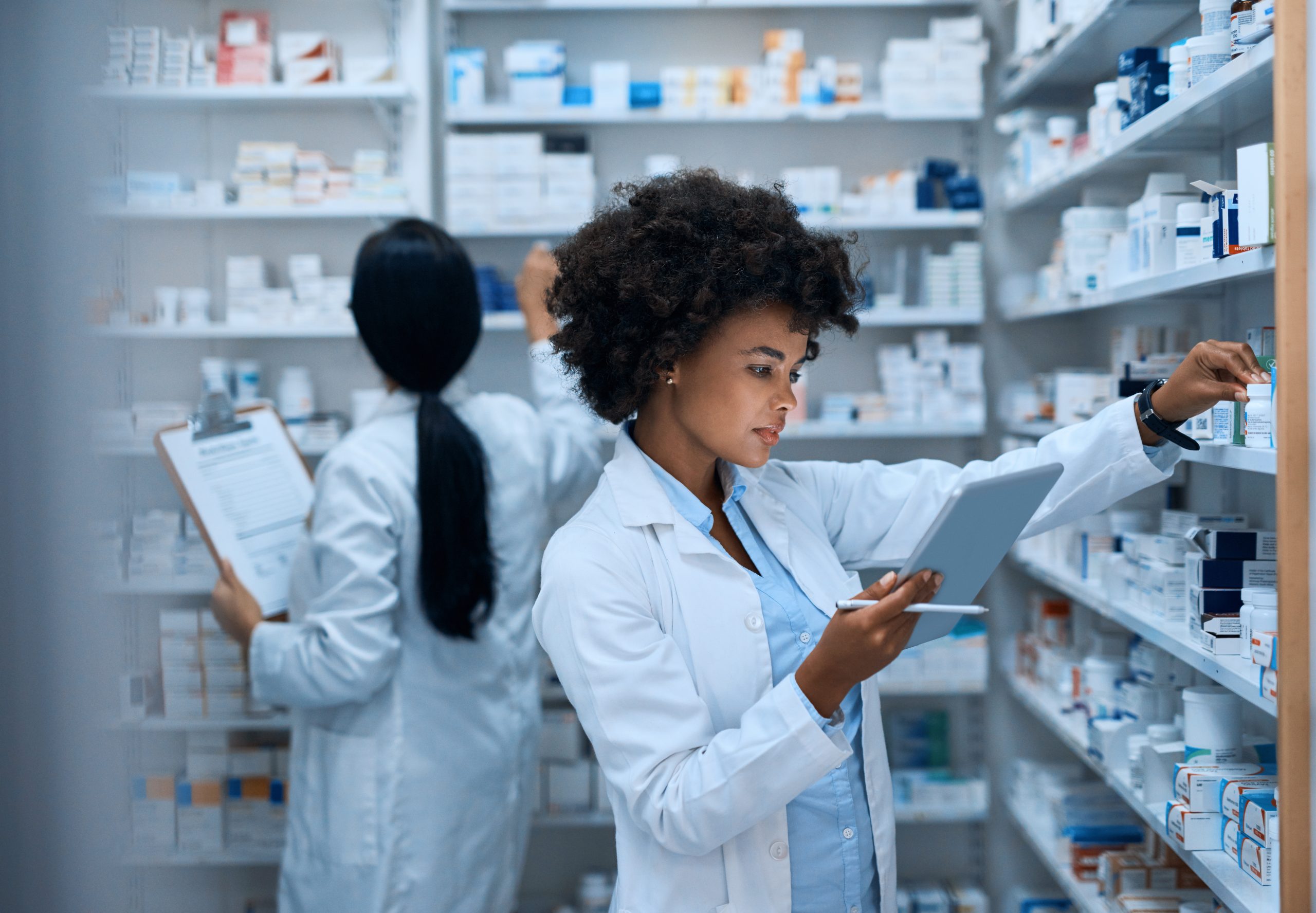 Two pharmacists are holding clipboards while checking shelves for prescriptions