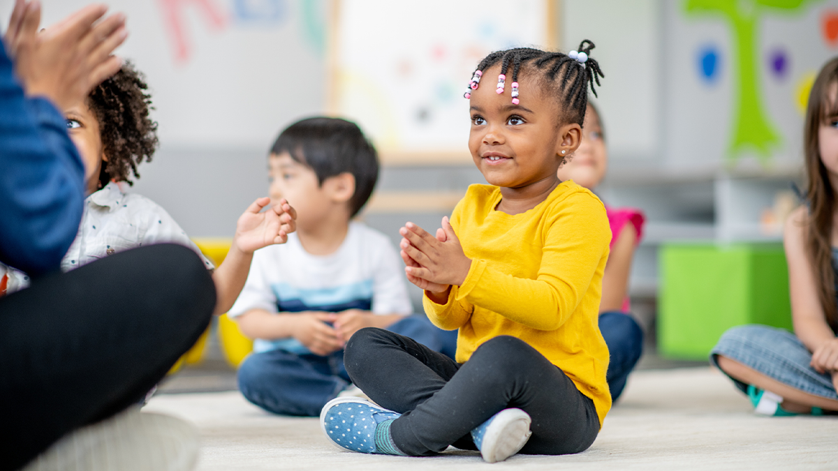 2 year old sits on a classroom floor clapping hands together.