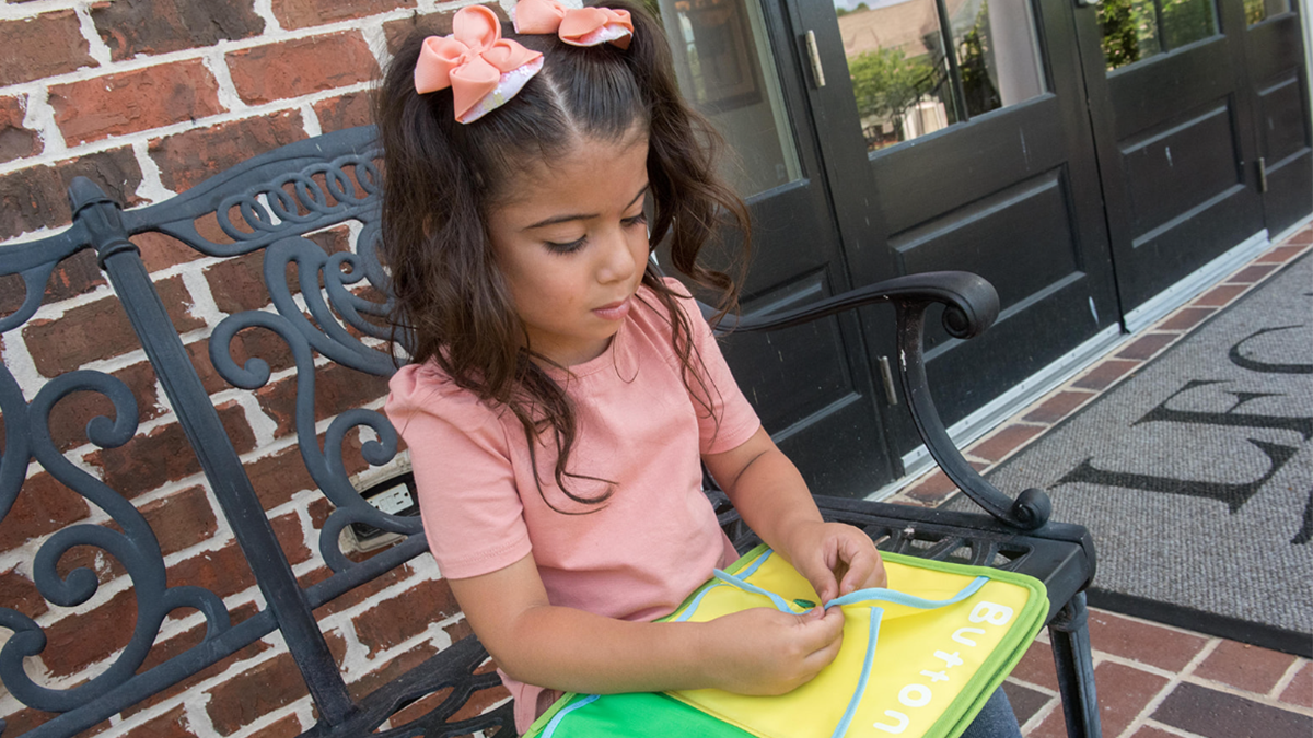 Una ni&ntilde;a peque&ntilde;a sentada en una banca abrochando dos trozos de tela en un libro.