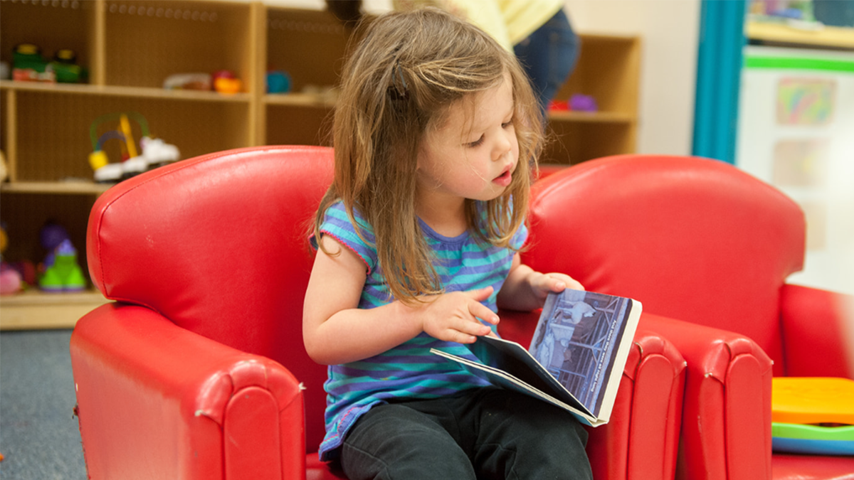 Una ni&ntilde;a peque&ntilde;a sentada en una silla hojeando un libro de ilustraciones.