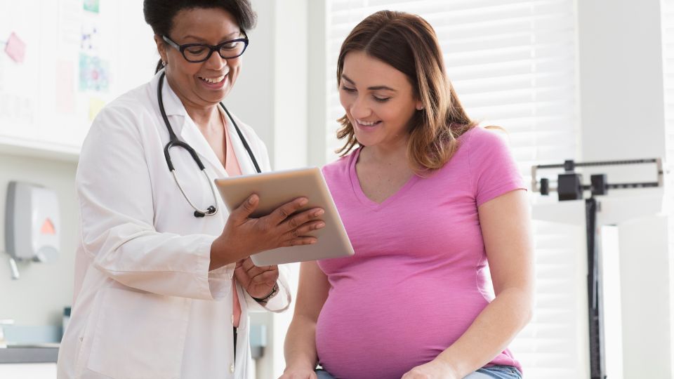Pregnant woman and doctor smiling, looking at a tablet in a healthcare office.