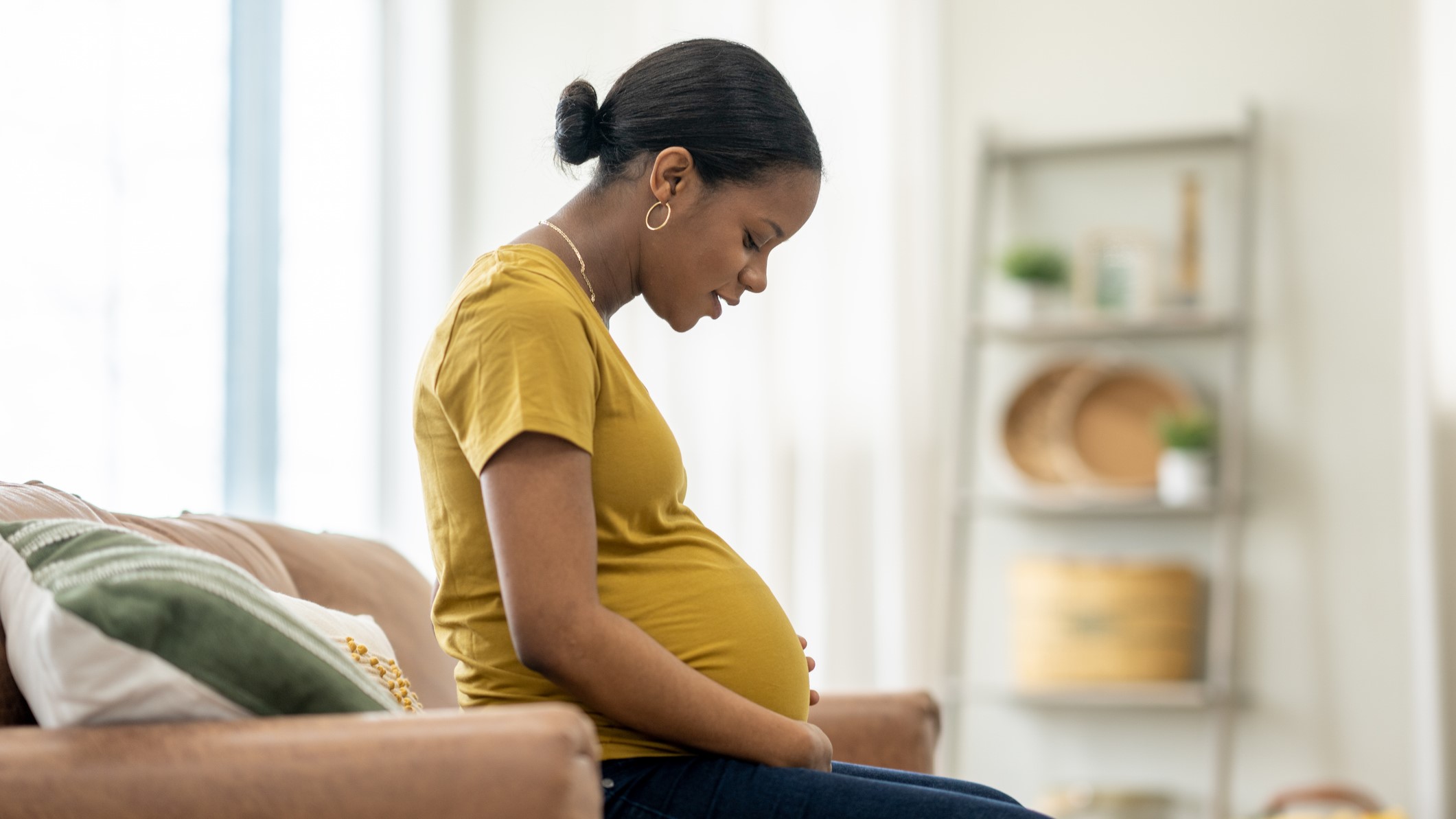 Pregnant woman in a yellow shirt sitting on a couch looking at her stomach.