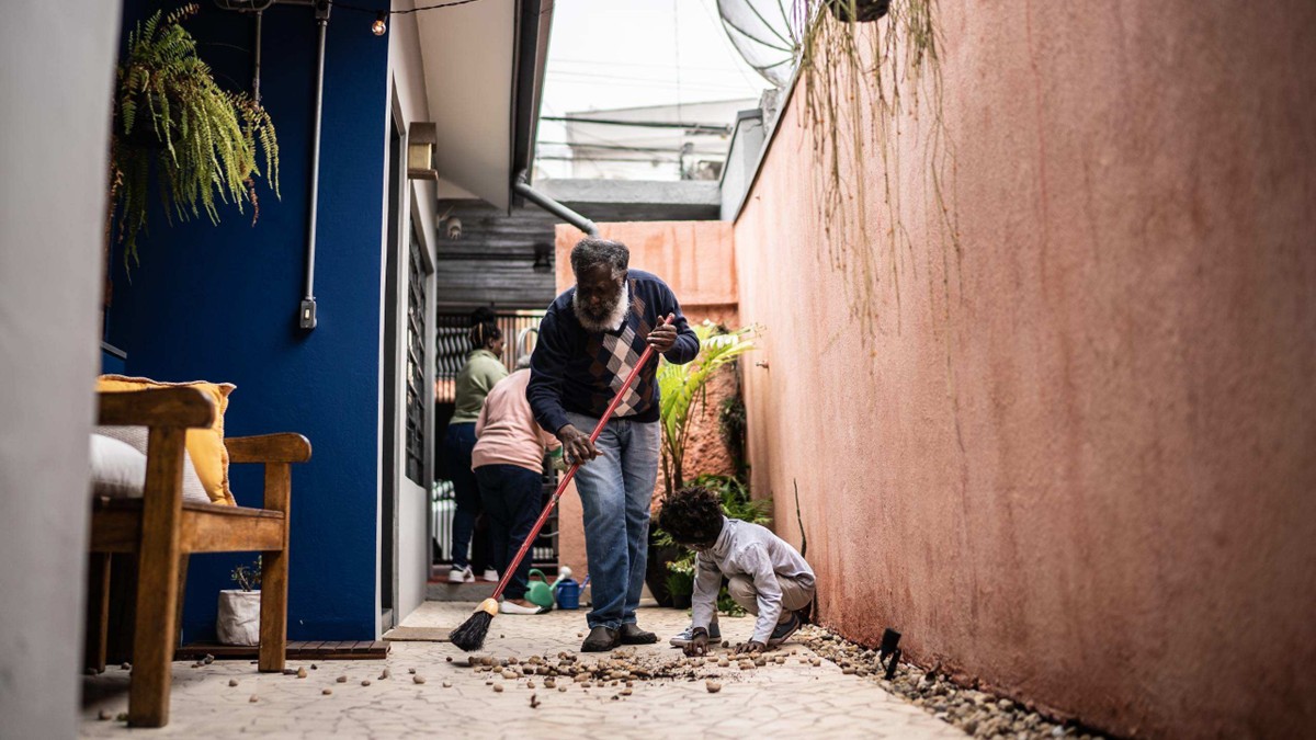 An elderly man and grandson sweep the floor of a rural house.