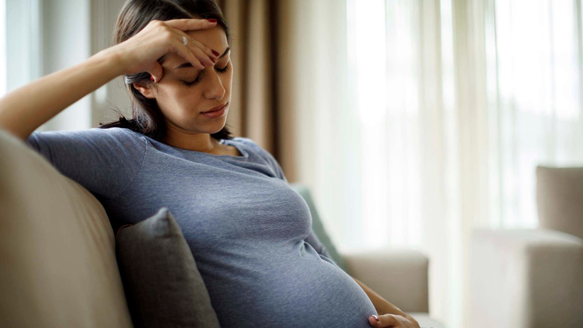 An ill pregnant woman sits on a couch holding her hand to her head.