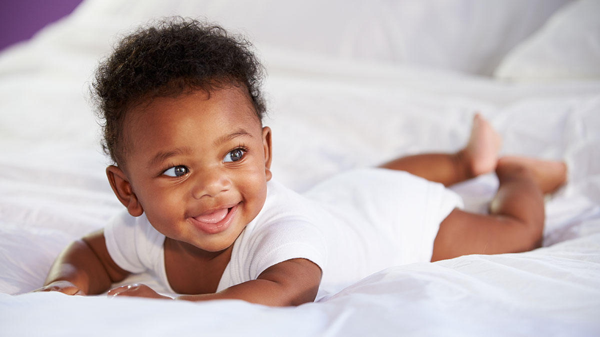 Smiling baby lying on their tummy on a white bed, looking off to the side.