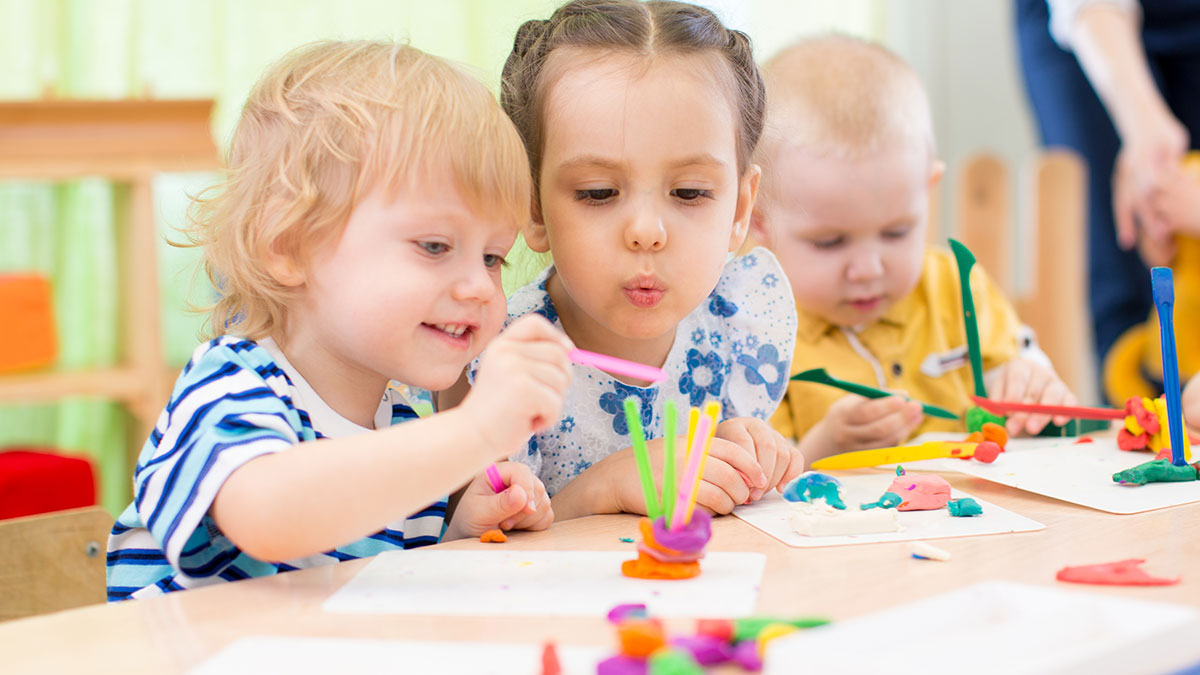Three toddlers at a table creating colorful crafts with clay and small tools.