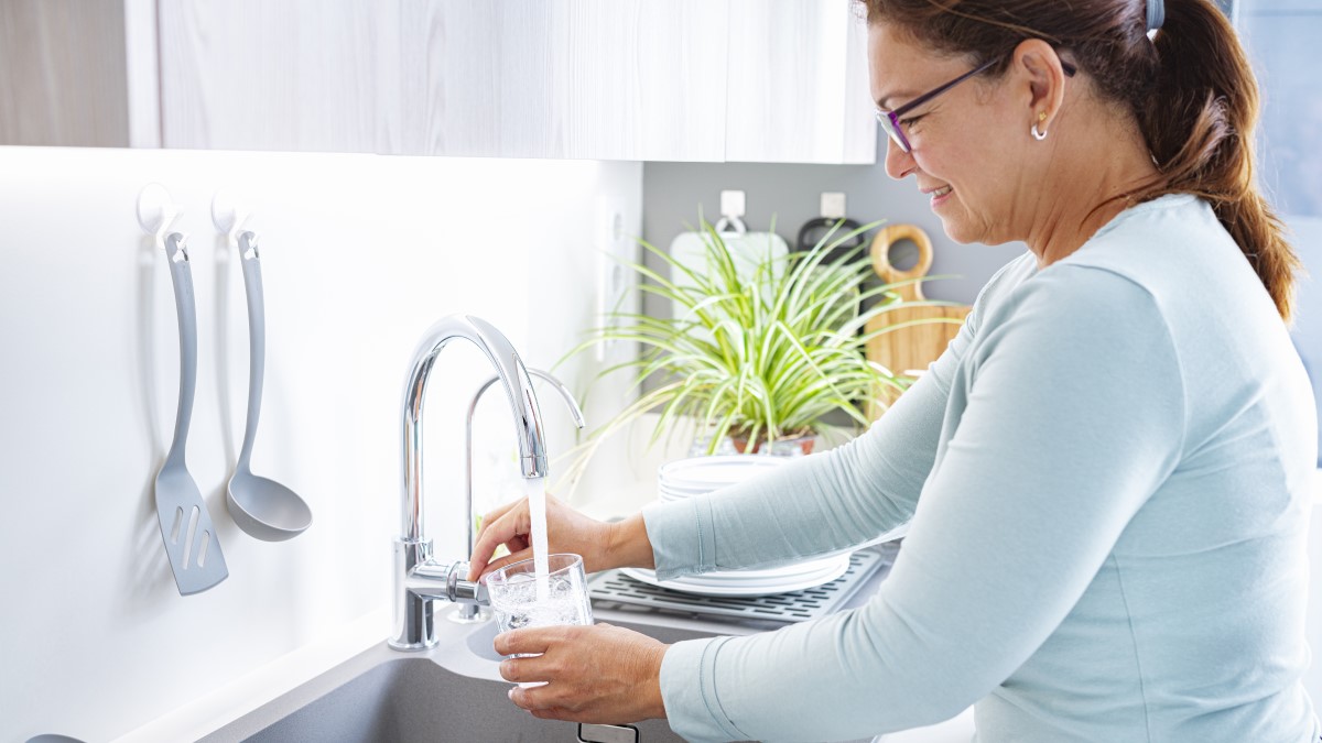 Mujer sonriendo y llenando un vaso con agua de la llave de la cocina.