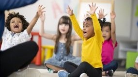 — title missing — Young children sitting on floor and raising hands in classroom.