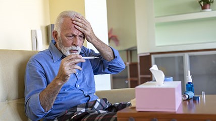 An older man holds hand to head and views thermometer.