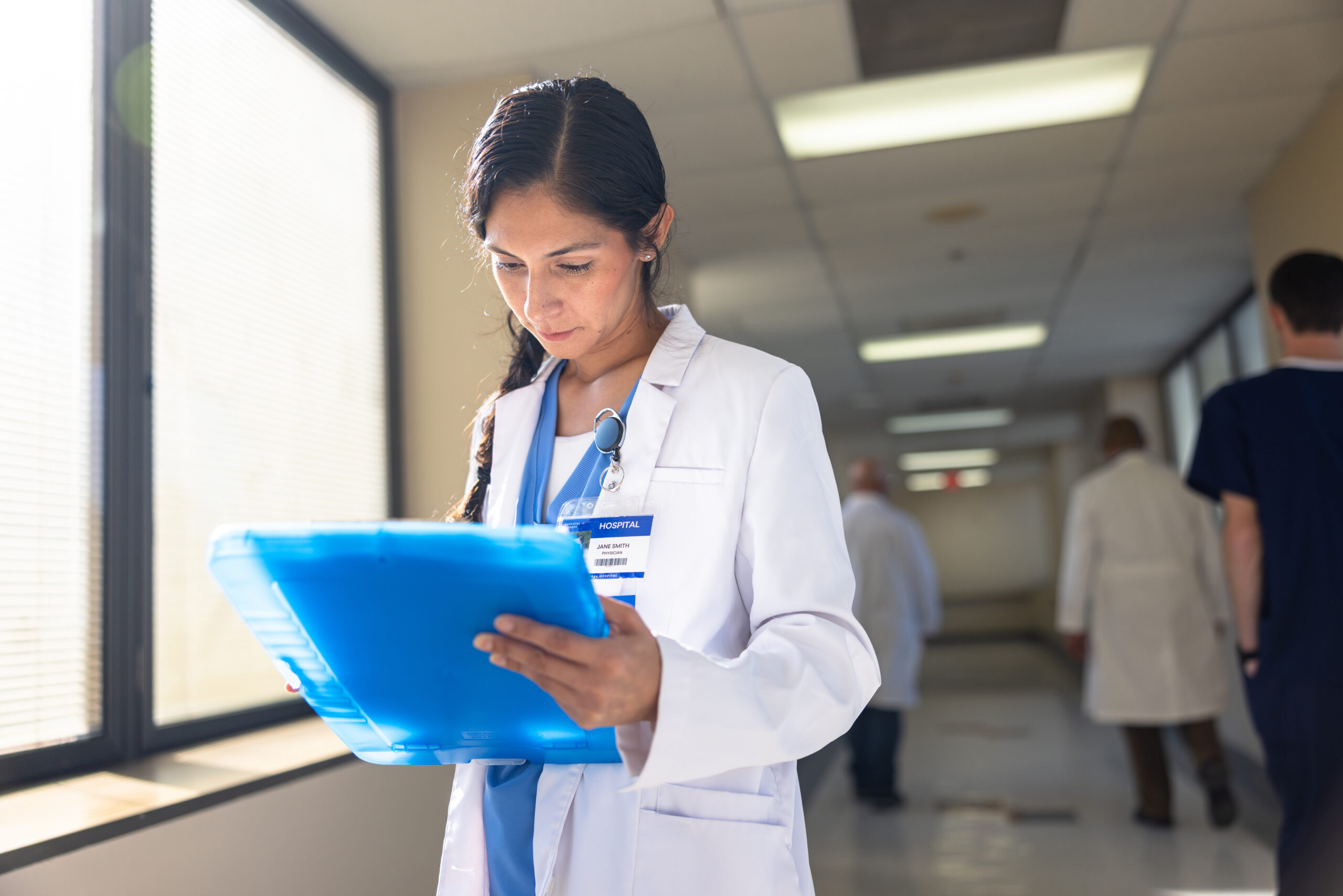 A clinician stands in a busy hospital corridor reading a clipboard.