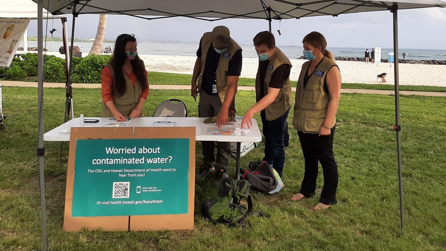 Four masked people stand under a white tent outside in Oahu. Sign in front says, "Worried about contaminated water?"