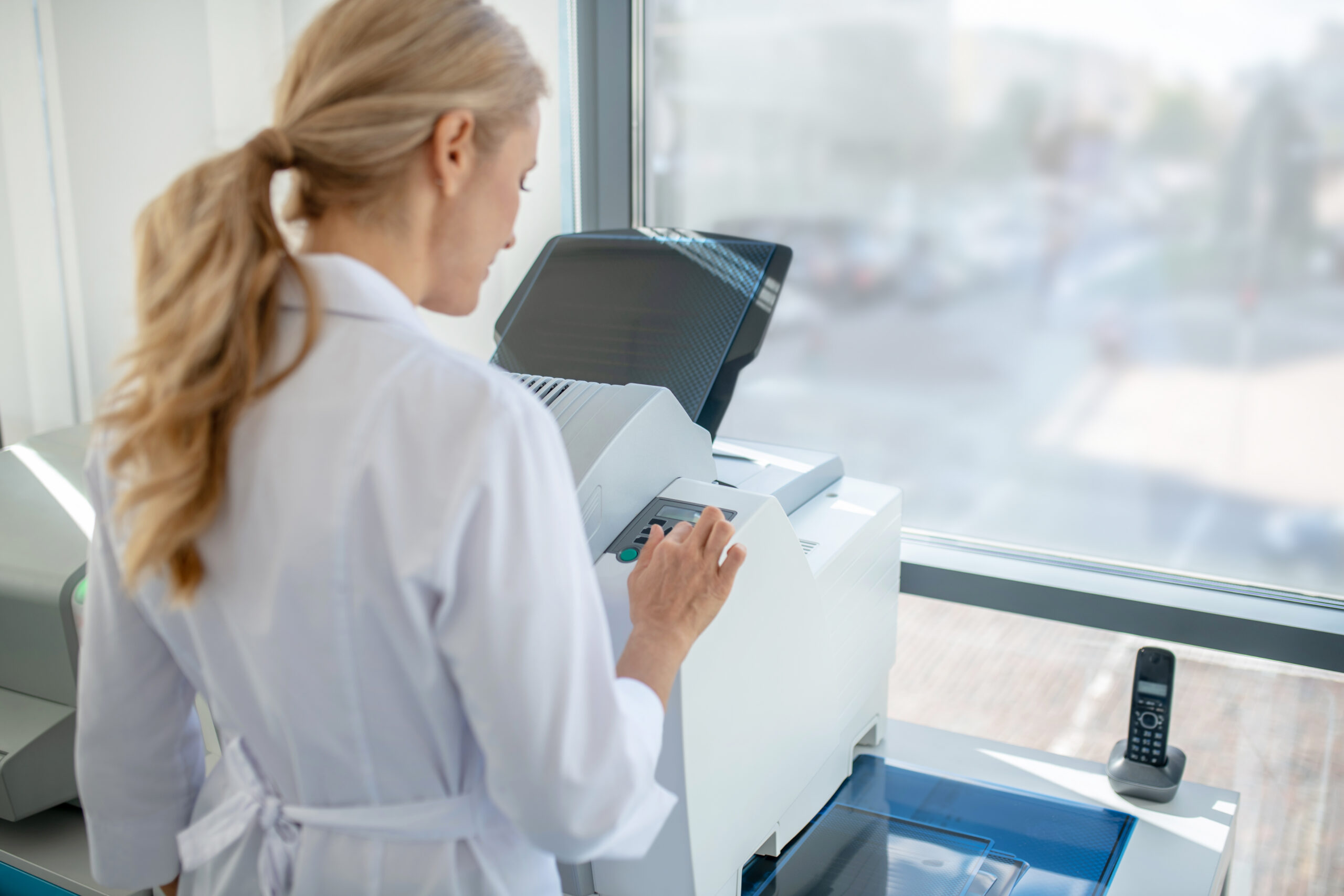Fair-haired young doctor having a busy day in clinic standing in front of and using a fax machine