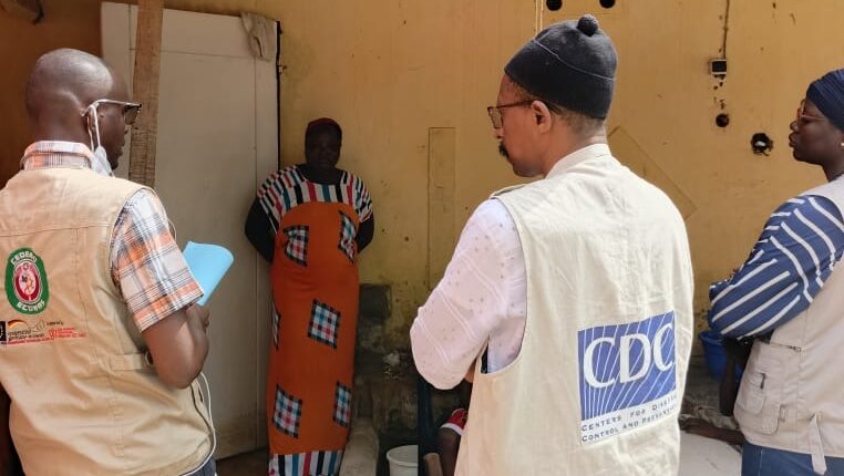 Public health team members speaking with a community member outside a small building during a field investigation.