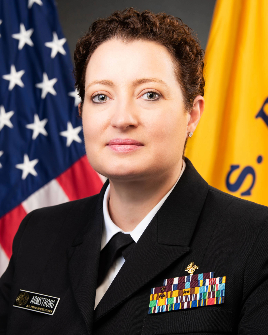 Portrait of Paige Armstrong, Director for CDC's Global Health Center, she wears a black Commissioned Corps uniform and stands against a background of the United States and Commissioned Corp flags.