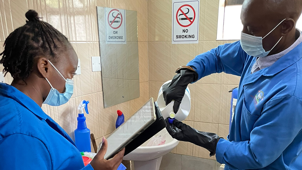 An evaluator wearing a surgical mask watches as a worker wearing a surgical mask who cleans toilets measures bleach.