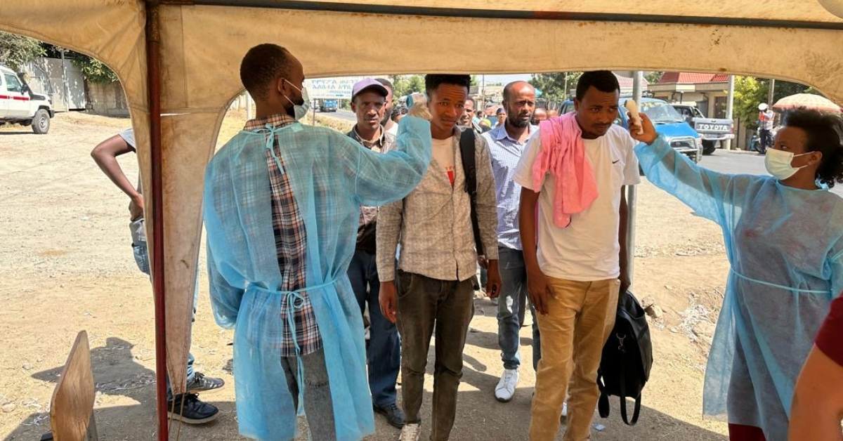 Two health workers in protective gowns and face masks screen a line of men by taking their temperatures outdoors.