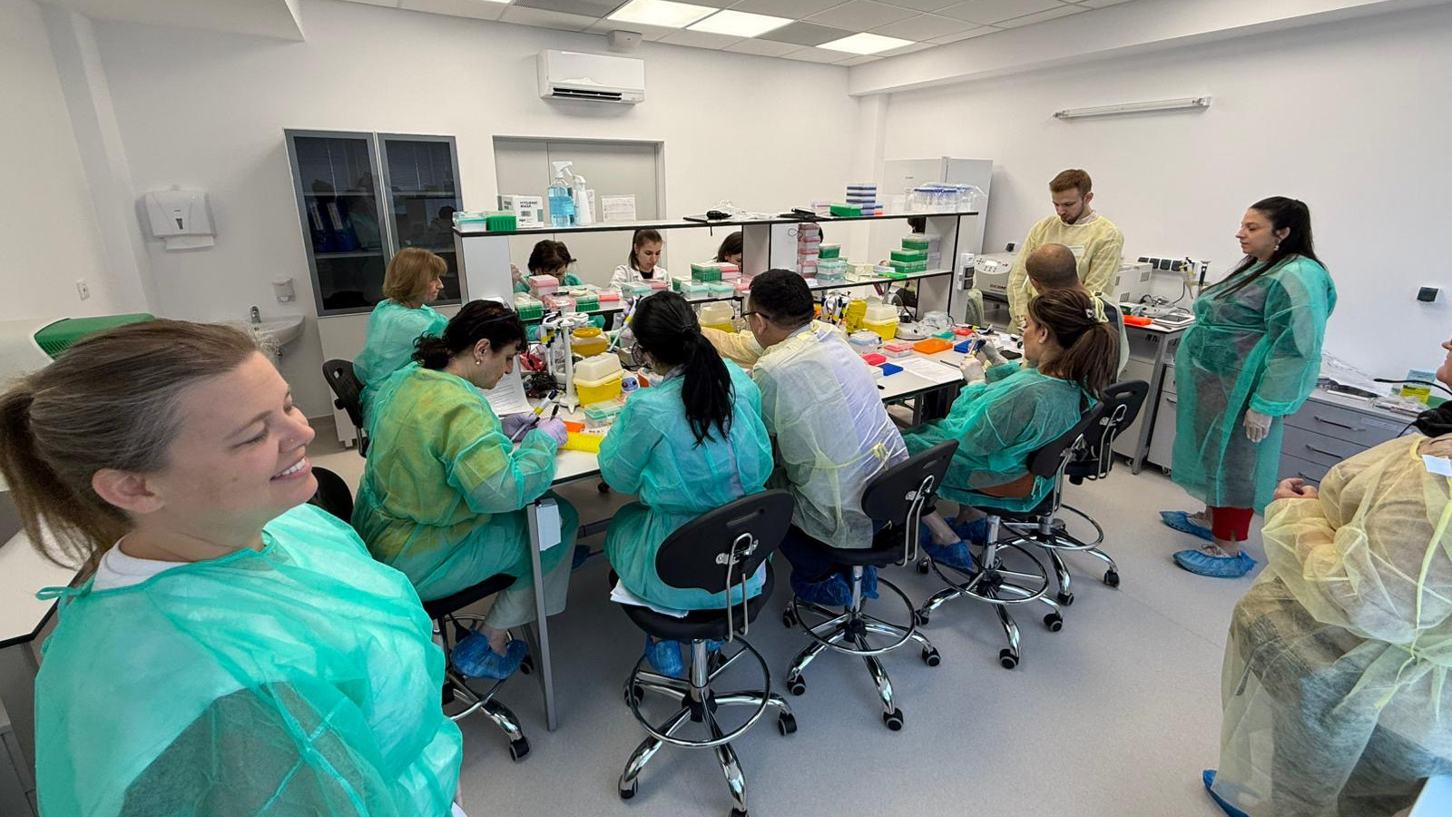 A group of people wearing green and yellow protective gowns and blue shoe covers are seated at a long table, using scientific equipment in a laboratory.