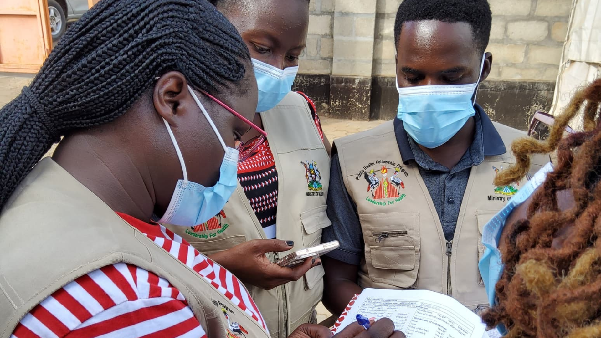 Four masked healthcare workers outdoors reviewing paperwork while one writes and another uses a phone.