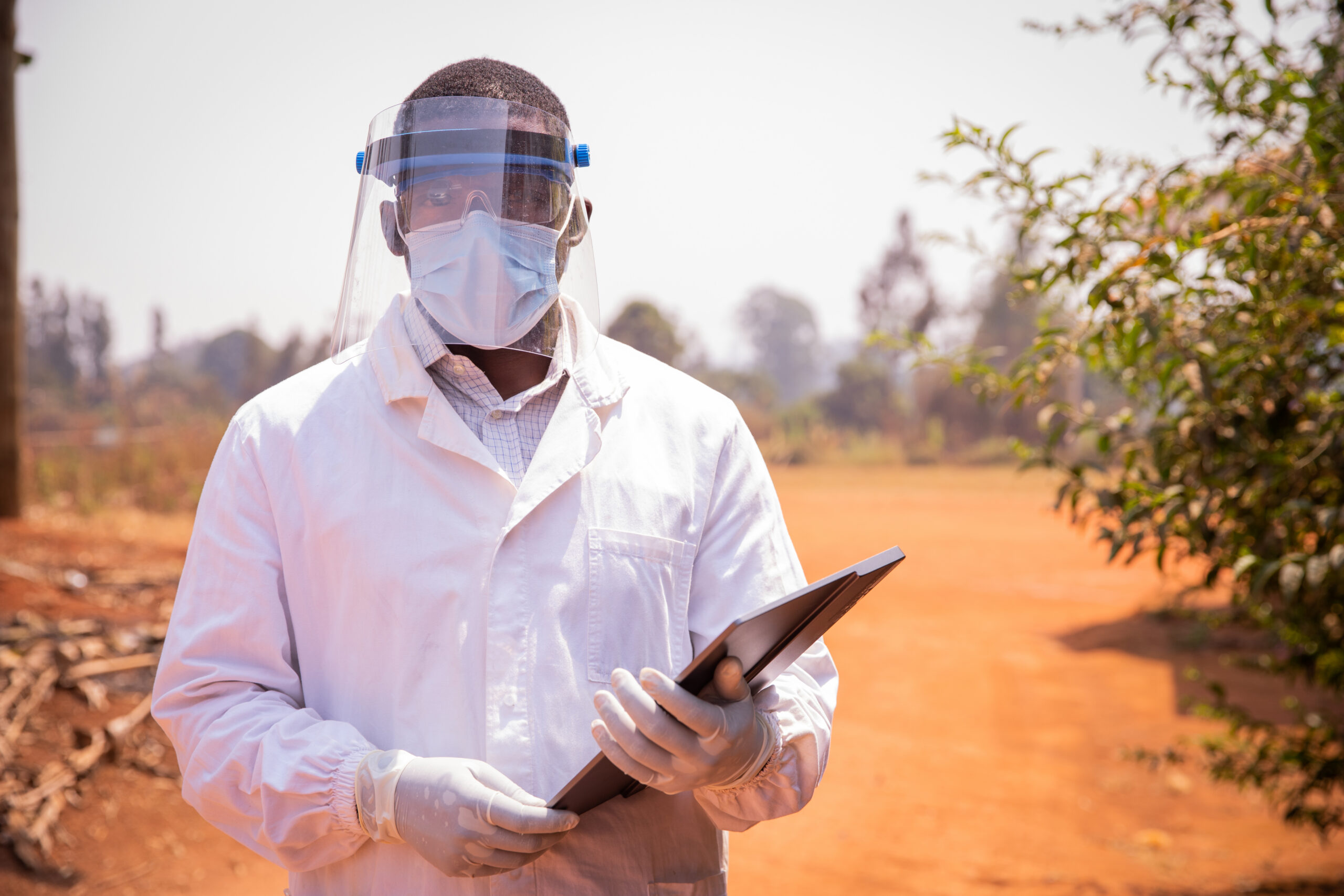 Image of a man in white coat and personal protective shield mask over face holds an ipad in the field.