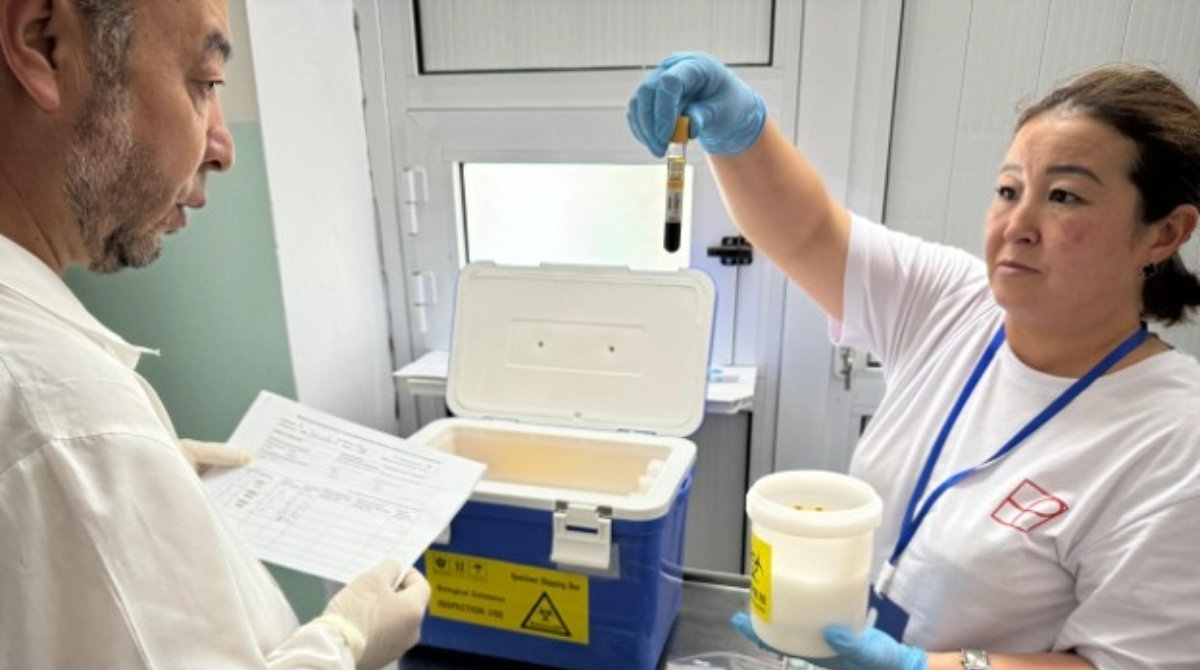 Two lab workers in gloves examine a blood sample vial and paperwork next to an open biohazard specimen transport cooler