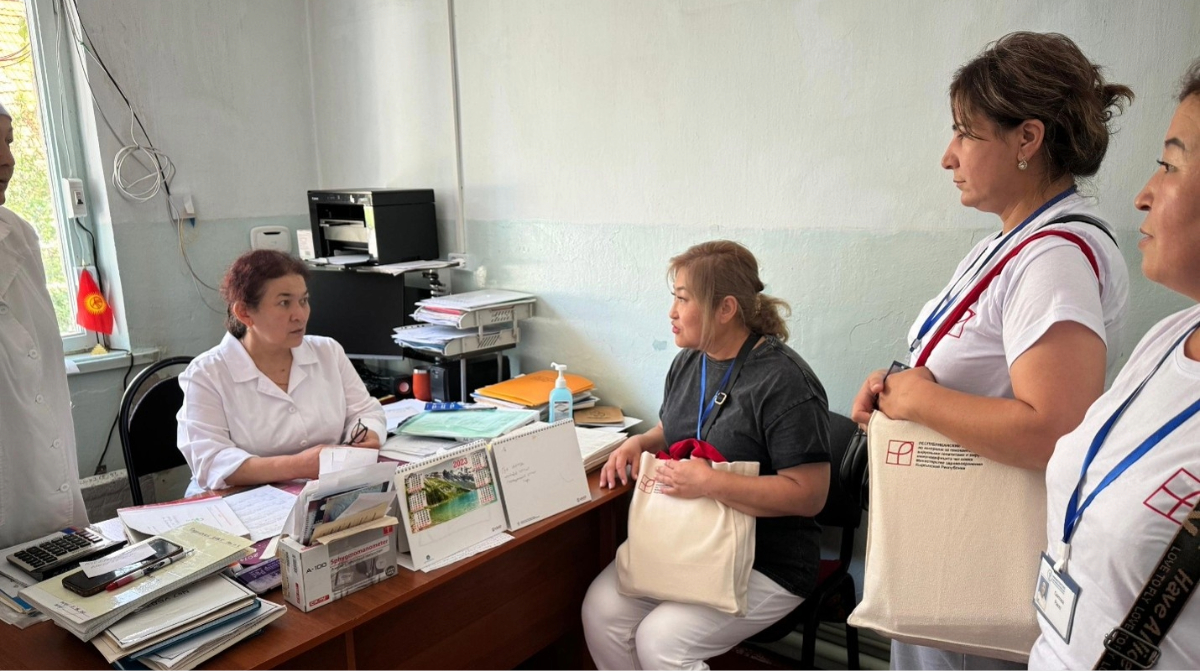 A healthcare worker in a white coat sits at a cluttered desk speaking with three visitors wearing lanyards in a clinical office setting.