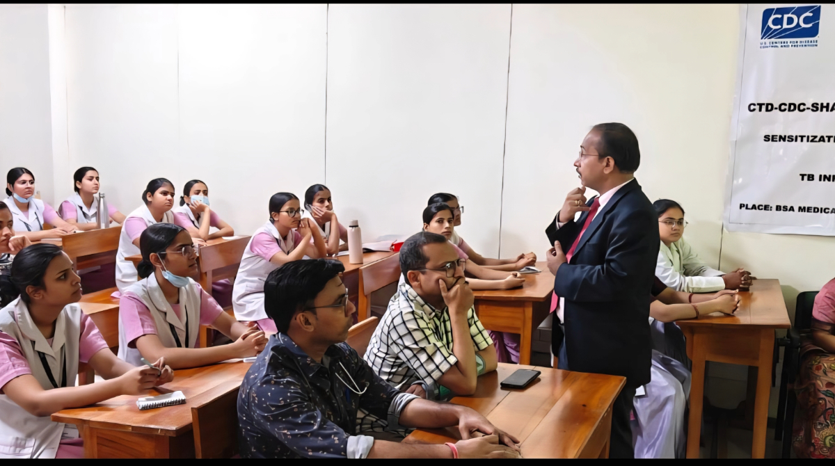 Man in a suit speaking to a classroom of nursing students and colleagues seated at desks, with a CDC banner for a TB infection sensitization training visible in the background.