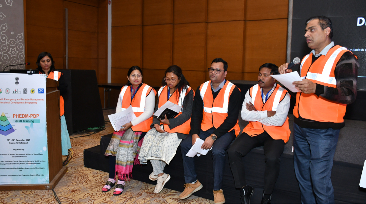 Man speaking into a microphone alongside four seated colleagues, all wearing orange safety vests, at the PHEDM-PDP Tier-III Training event.