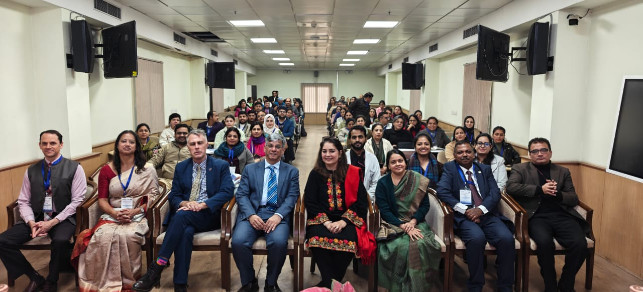 Group photo of conference attendees and dignitaries seated in a long meeting hall, with a row of officials in front and rows of attendees wearing lanyards seated behind them.
