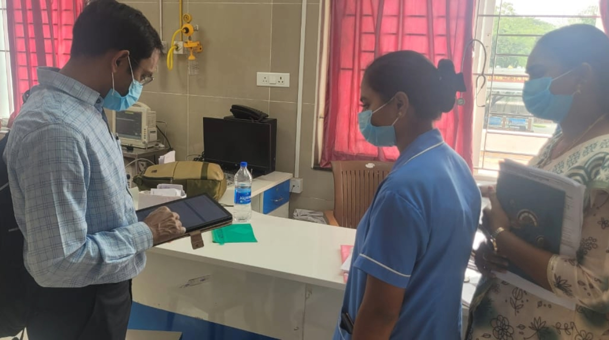 Man in blue shirt using a tablet at a clinic reception desk while a nurse in scrubs and a woman, both wearing face masks look at the man.
