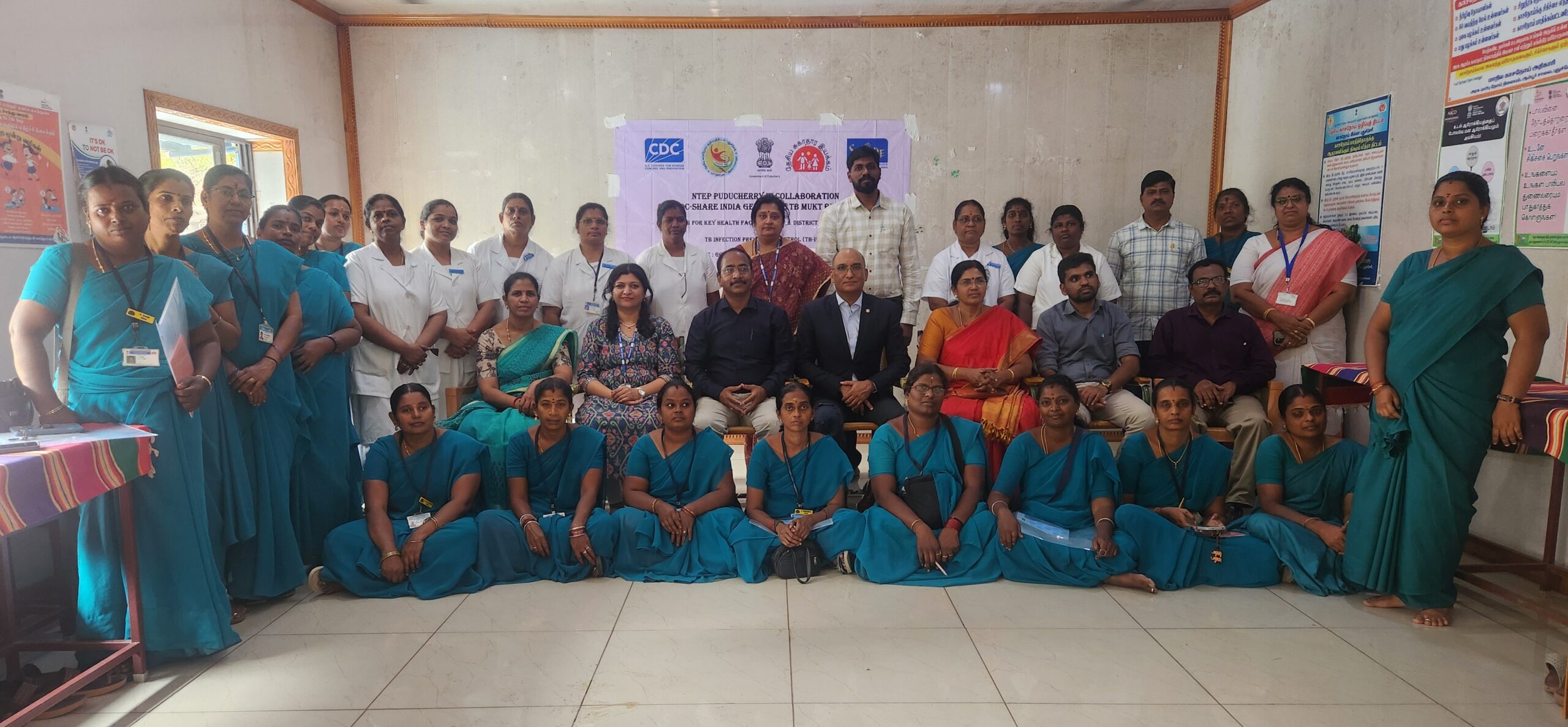 Group photo of approximately 30 people at an indoor training event, including seated officials in business attire, medical staff in white coats, and community health workers in teal uniforms with lanyards, posing in rows in front of a banner reading 'NTEP Puducherry in collaboration with C-SHARE India' about TB infection prevention and control training, with CDC and Government of India logos visible.
