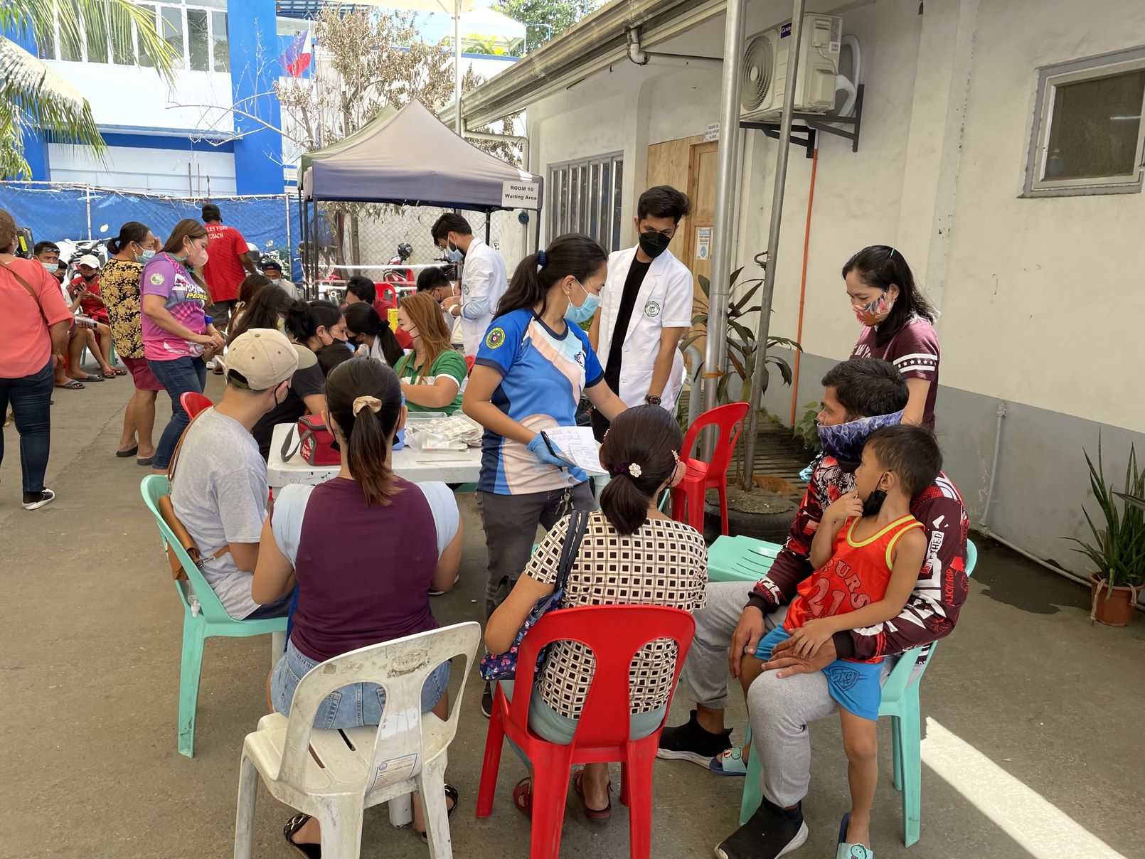 Many people with face masks covering their nose and mouth are standing or sitting outdoors near a table and tent.