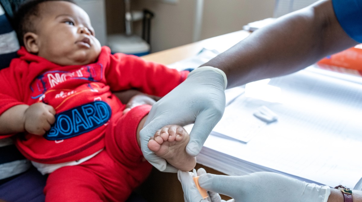 A baby in a red outfit gets a heel prick test from a gloved medical worker at a clinic.