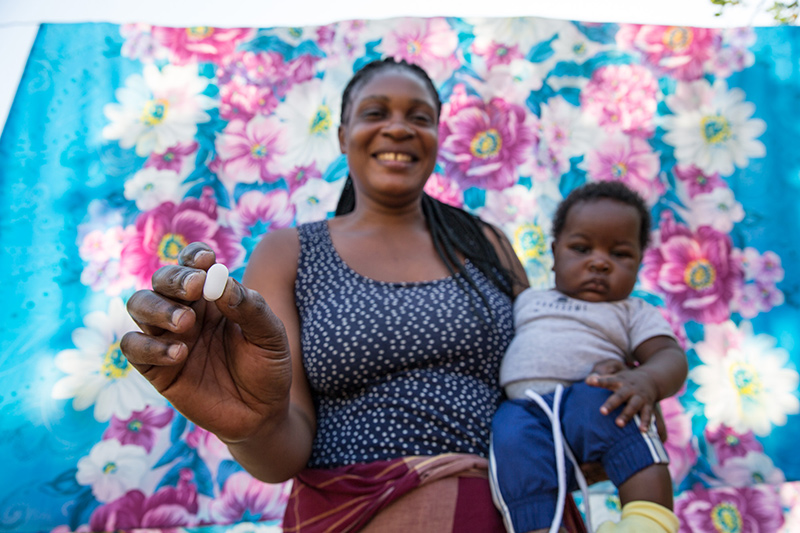 A smiling woman holds up a white pill while seated with a baby on her lap, in front of a colorful floral backdrop.