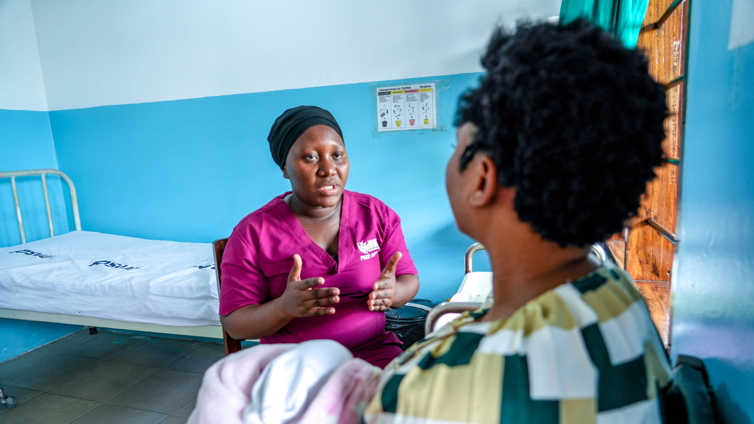 A health worker in a magenta scrub top gestures while talking with a patient in a clinic room.