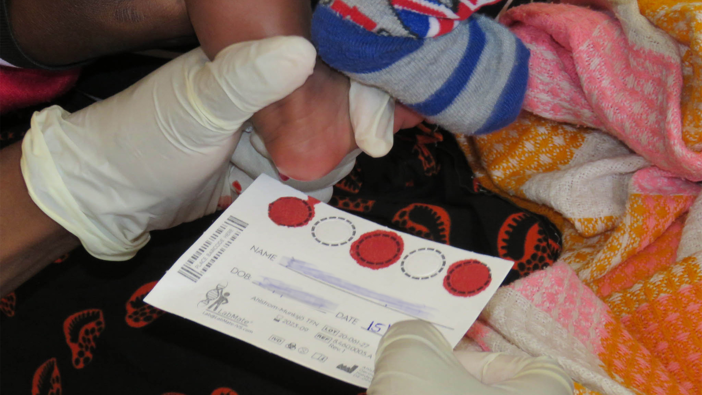 A gloved healthcare worker holds a newborn baby's heel while filling in blood spots on a card.