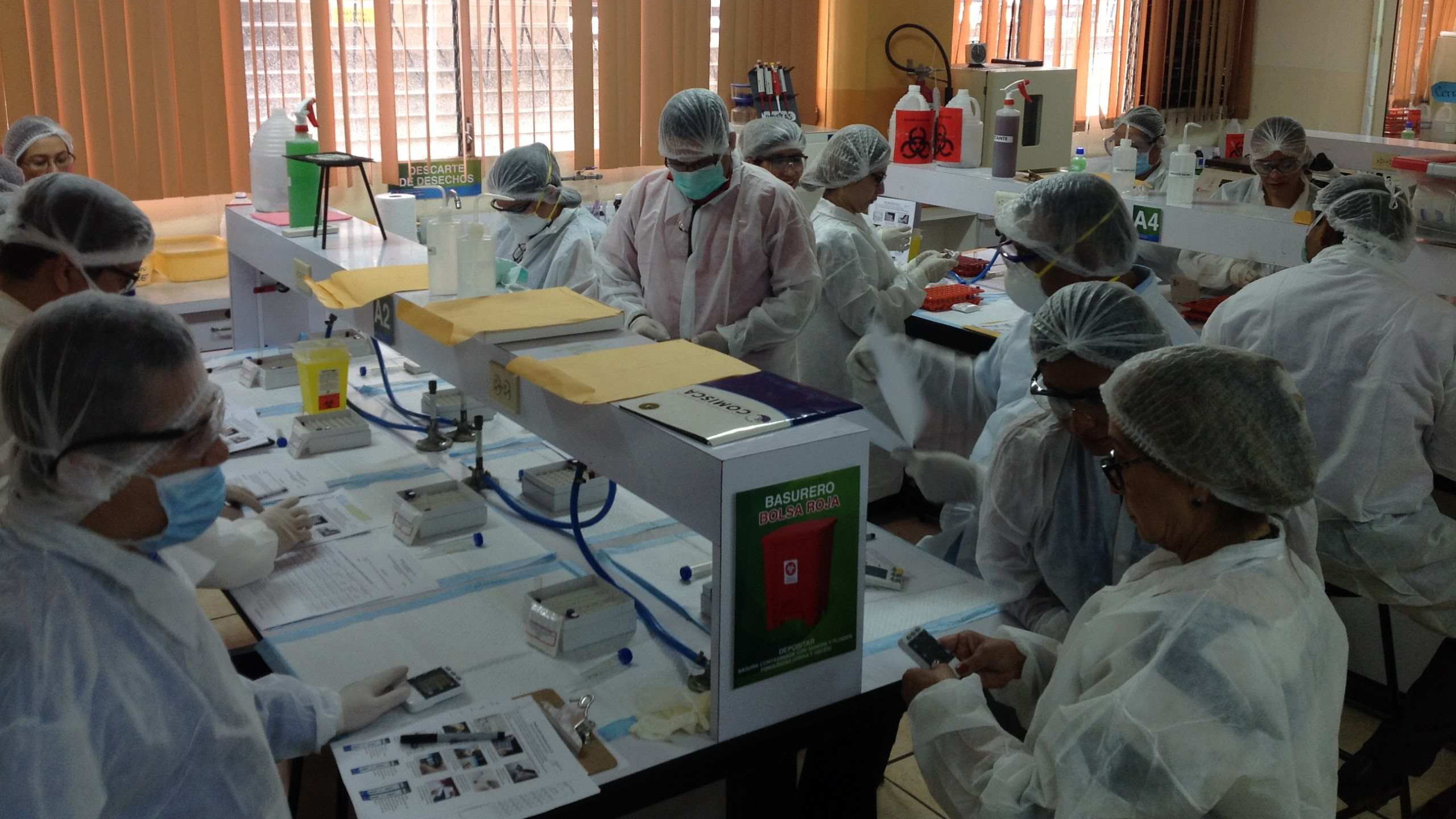 Lab technicians in protective gear process samples at shared testing workstations.