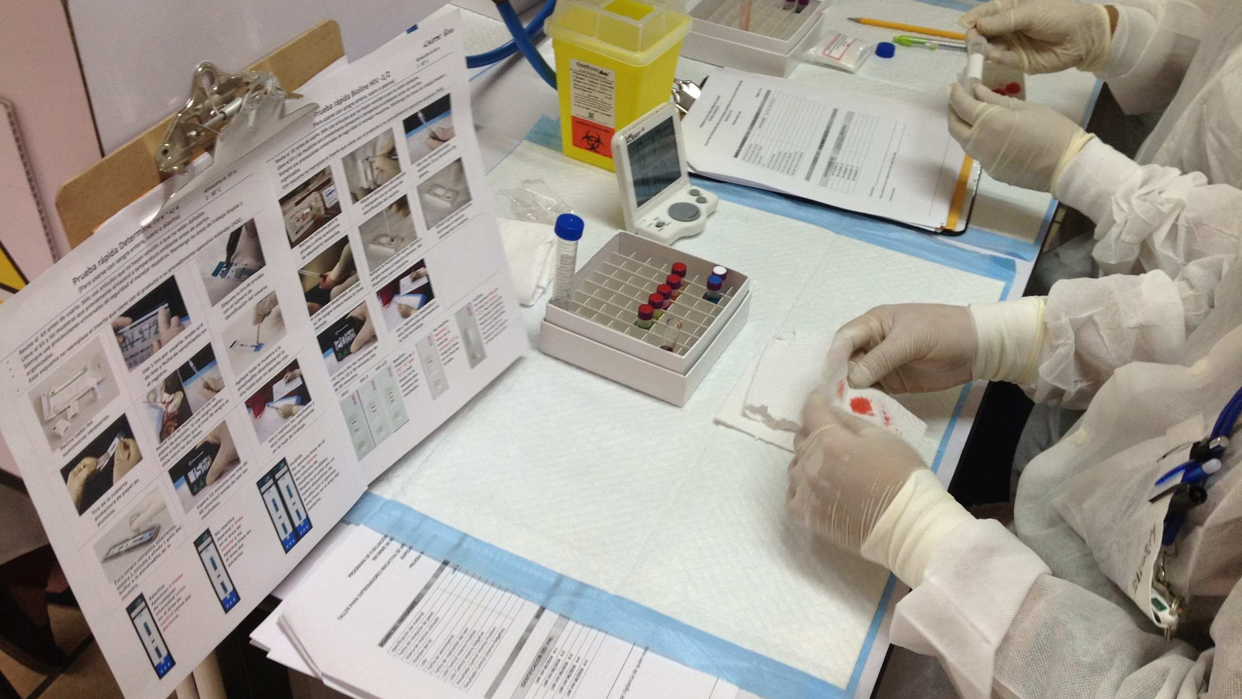 Lab technicians in protective gear process samples at shared testing workstations.