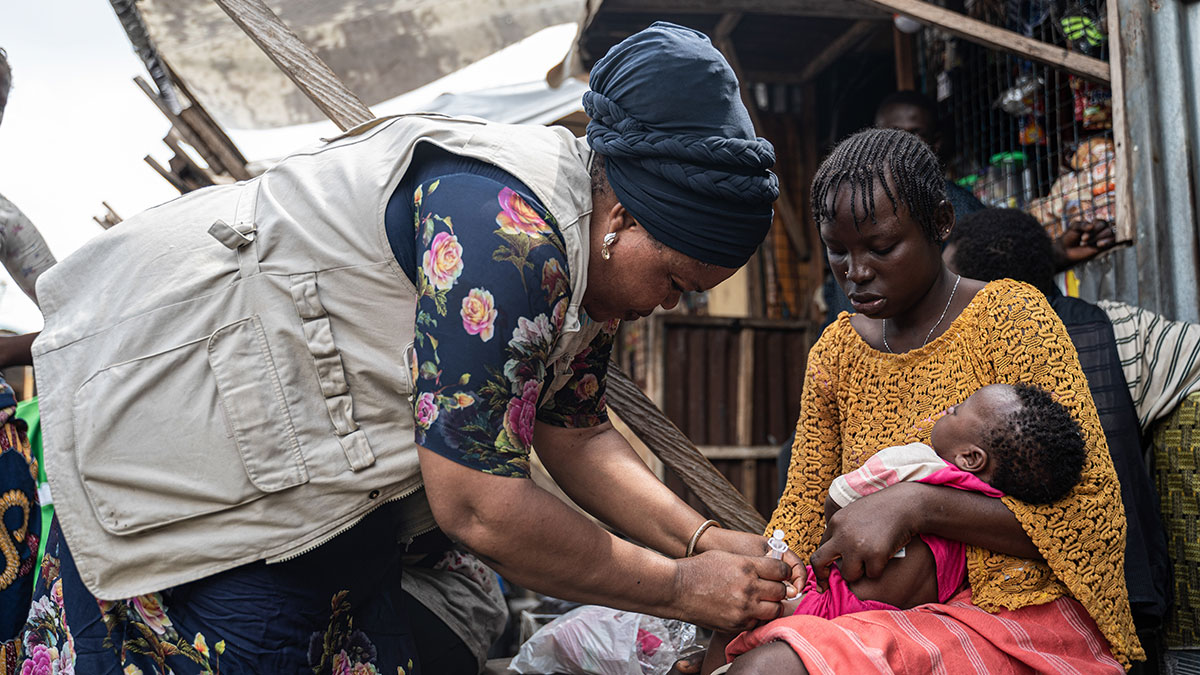 A woman wearing a khaki vest gives an infant a shot in her leg while the mother holds the baby.