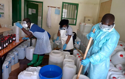 Three workers wearing masks are surrounded by large containers of alcohol-based hand rub