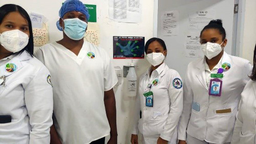 Three female and one male healthcare workers wearing masks stand in front of a dispenser of alcohol-based hand rub and a poster promoting hand hygiene