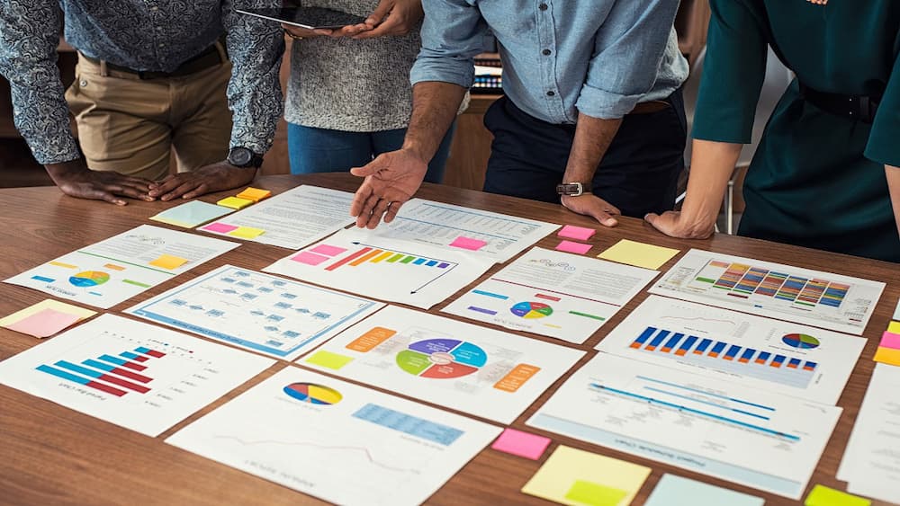 Printed paper with different graphs and charts laid out on a table, with coworkers standing around the table.