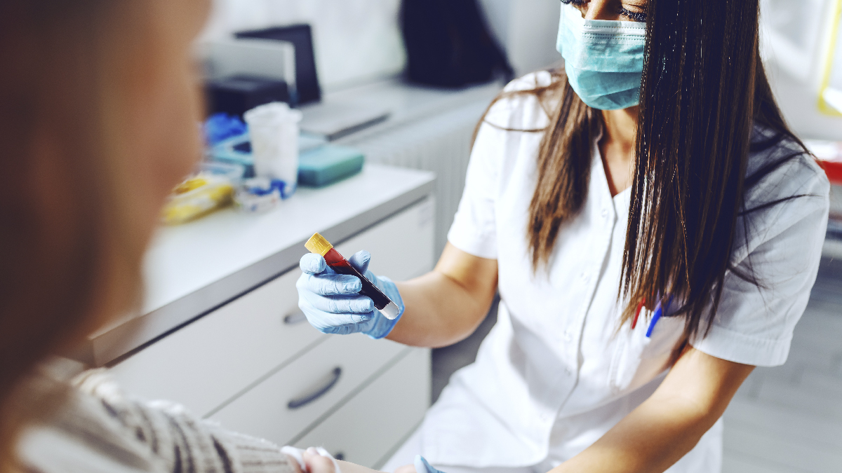 Mujer en un entorno médico, con un uniforme médico blanco y mascarilla, sosteniendo un vial con sangre.