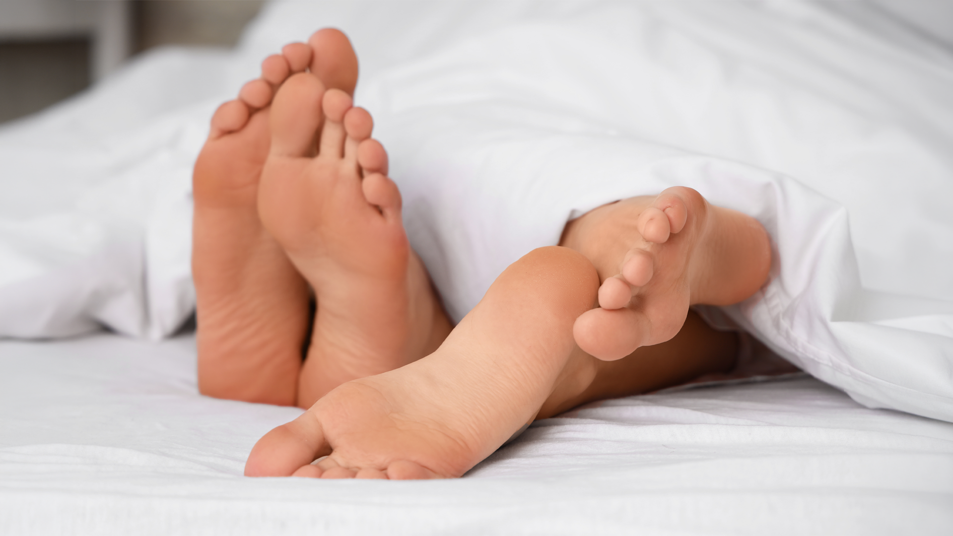 Dos pares de pies asom&aacute;ndose por debajo de las s&aacute;banas de una cama.
