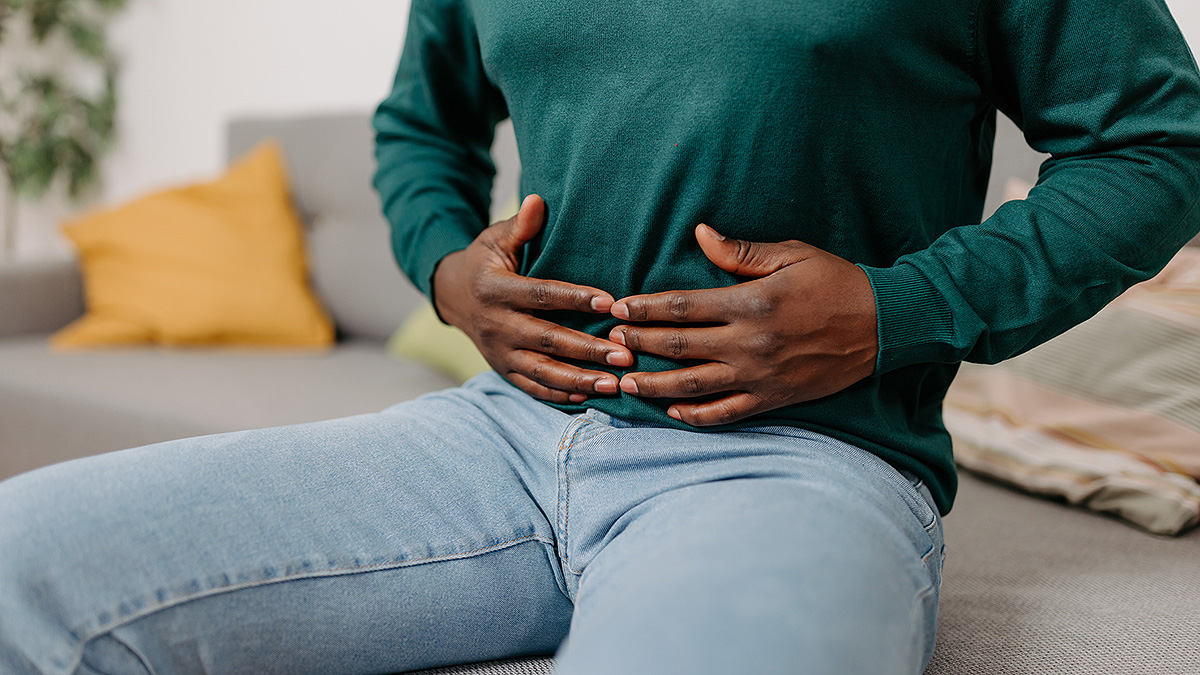 ibd man with stomach pain A young African American man sits on a sofa, holding his stomach from digestive pain.