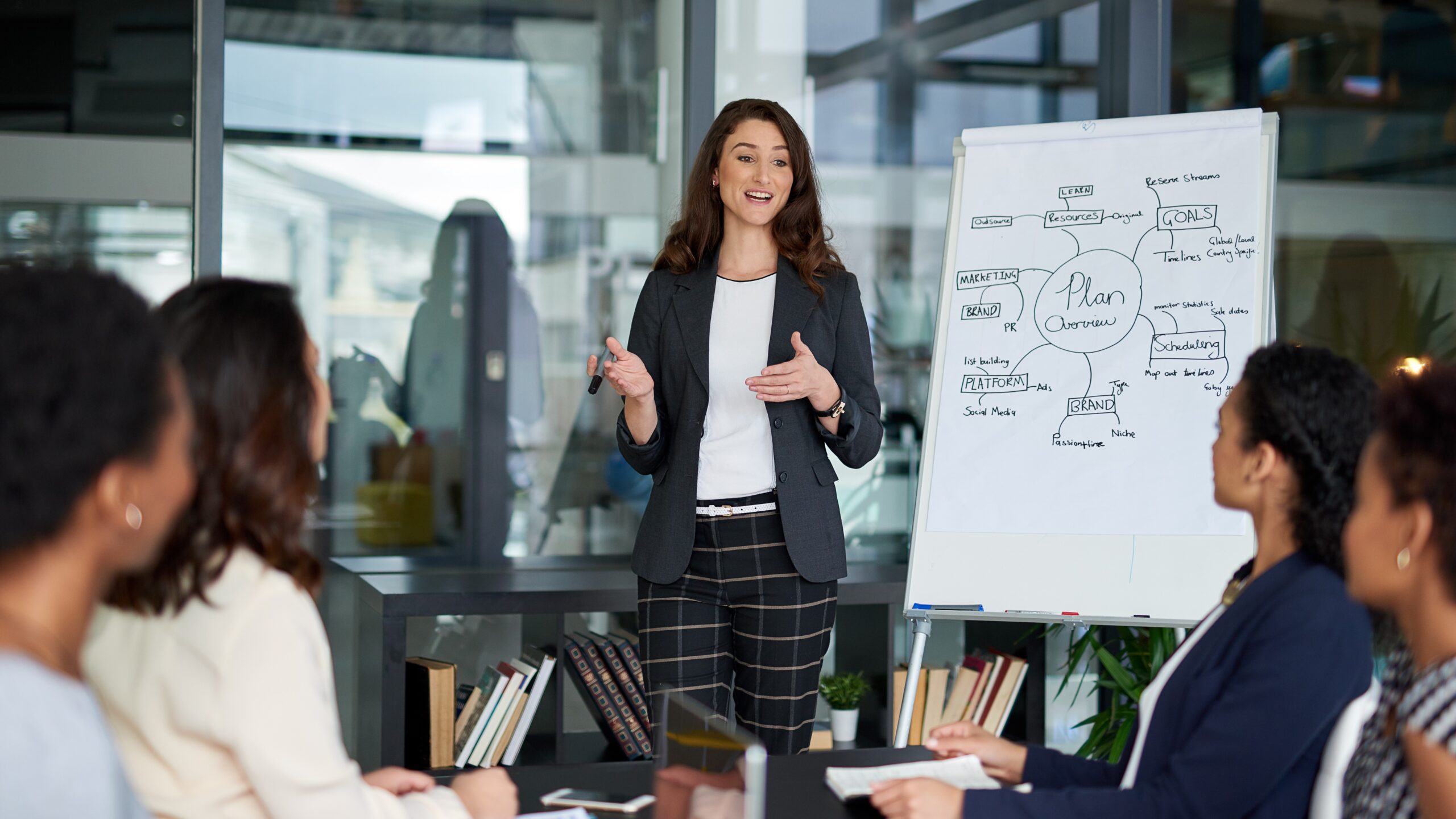 Staff sitting around a table having a discussion with someone standing at a paper flip chart with text on it.