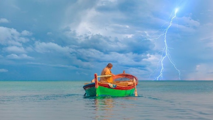 A man in a boat in the ocean with lightning in the background.