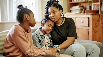 Una madre joven consuela a sus dos hijas pequeñas mientras están sentadas en un sofá.
