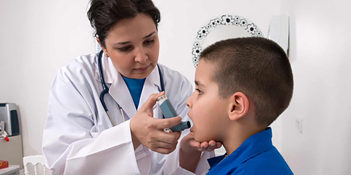 Image depicting a doctor preparing to use an inhaler on a young, asthmatic boy. The doctor&rsquo;s hand is on the child&rsquo;s chin and the child&rsquo;s mouth is open to use the inhaler.