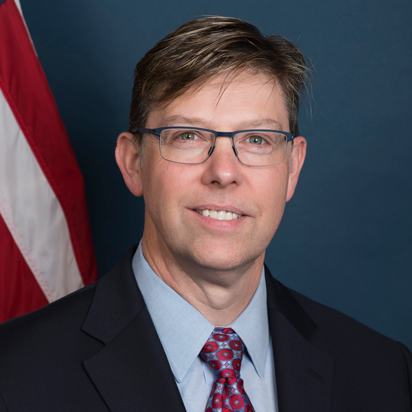 Portrait of a white man with blond hair and glasses, in a blue suit, blue shirt, and red tie, in front of a US flag