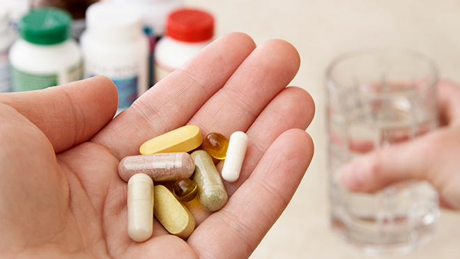 A man’s hand holding various dietary supplement pills and vitamins with a glass of water in the other hand
